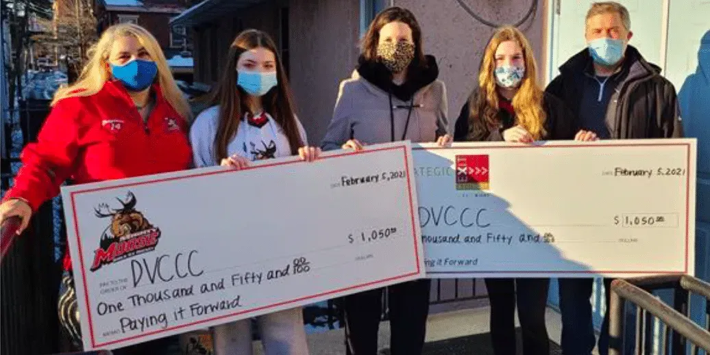 Five masked people holding large checks, presumably a donation, outside a building.