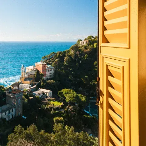 View of coastal village from yellow shuttered window, blue sea, green hillside.