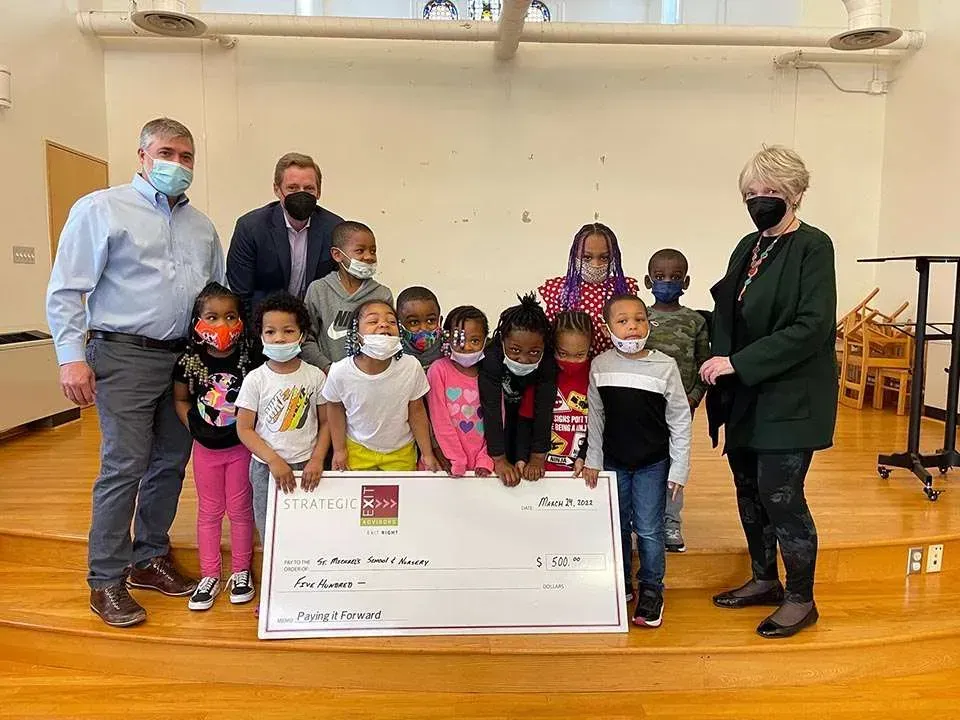 People presenting a large check to a group of children in a brightly lit room.