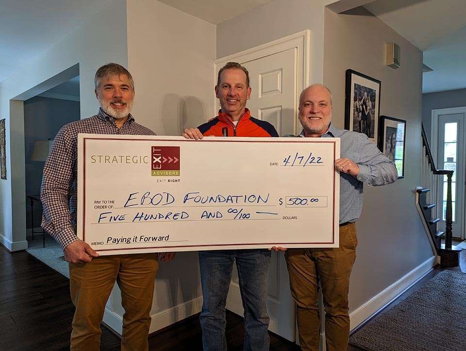 Three men holding a giant check for the EBOD Foundation, inside a home.
