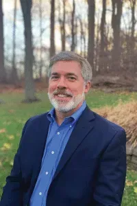 Man with graying hair and beard in a blue suit smiles outdoors with a forest background.