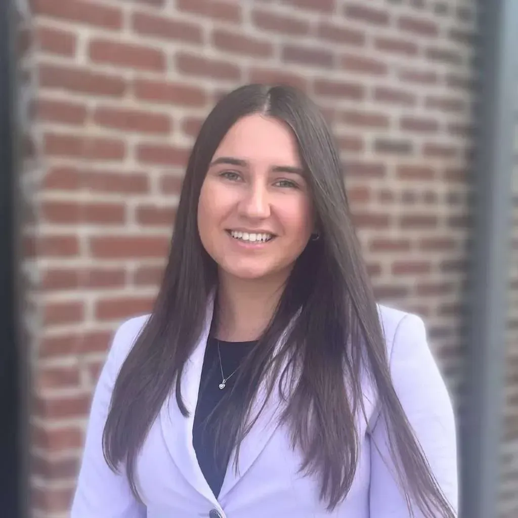 Woman with long dark hair smiles, wearing a light purple blazer, in front of a brick wall.