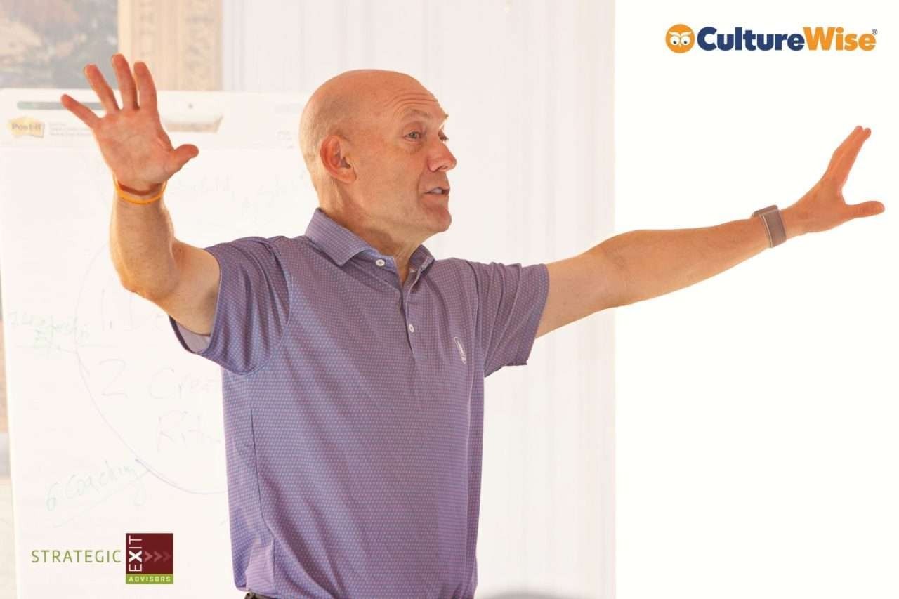 Man with arms outstretched, speaking.  He wears a purple polo shirt, standing in front of a whiteboard.