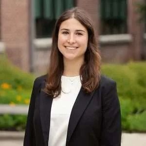Woman in a black blazer smiles, stands outdoors in front of a brick building.