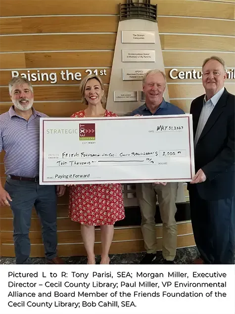 Four people hold a large check. Library setting with a lighthouse backdrop.