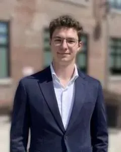 Man wearing glasses, a navy blazer, and a white shirt, smiling in front of a brick building.