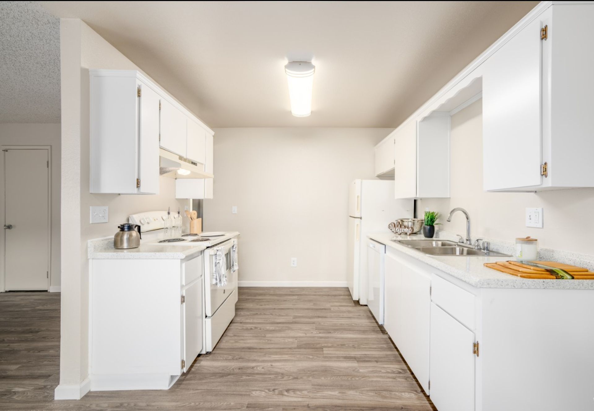 Bright, empty galley kitchen with white cabinets, white appliances, and wood-look flooring