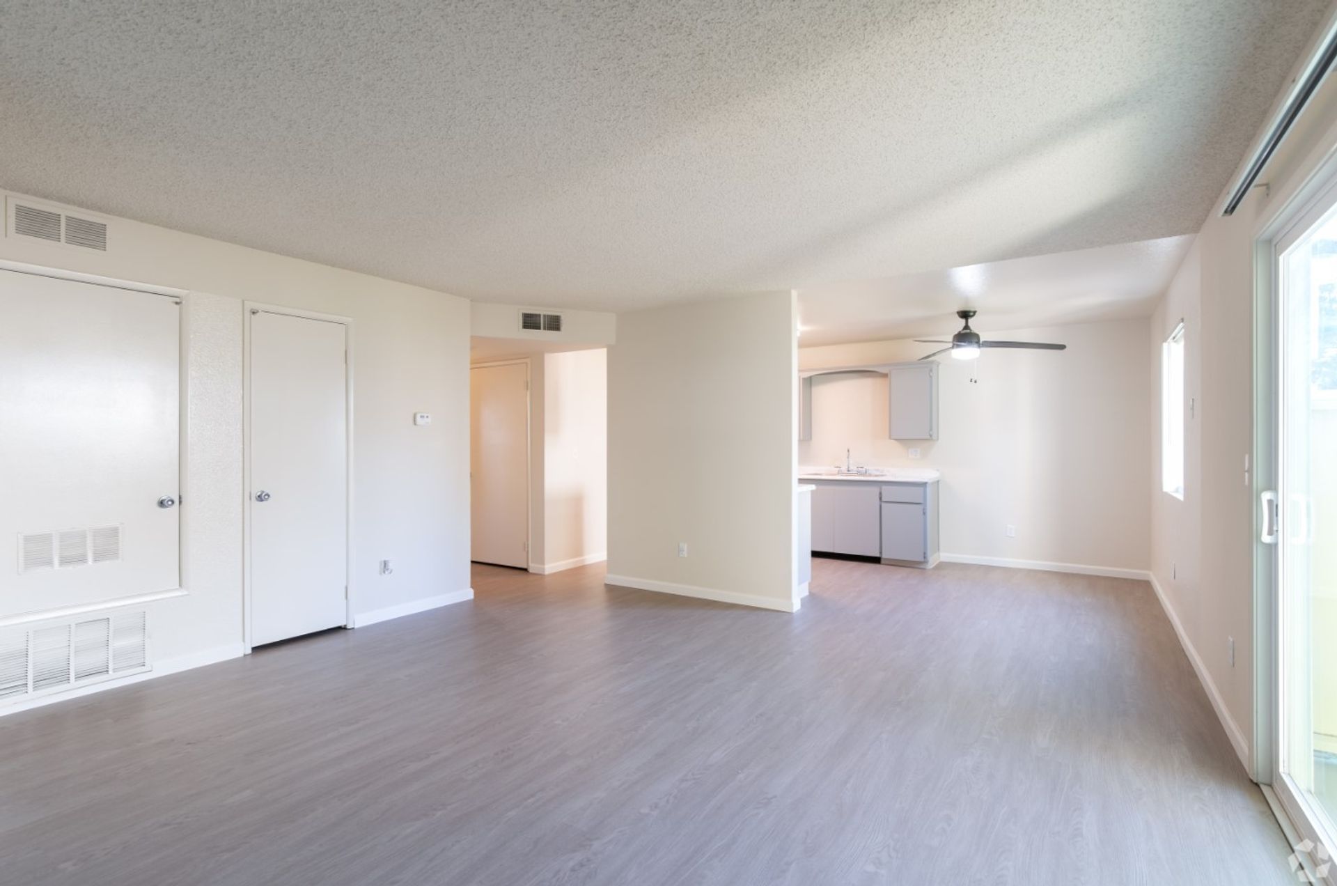 Empty modern apartment living room with light wood floors, white walls, and an open kitchen area.