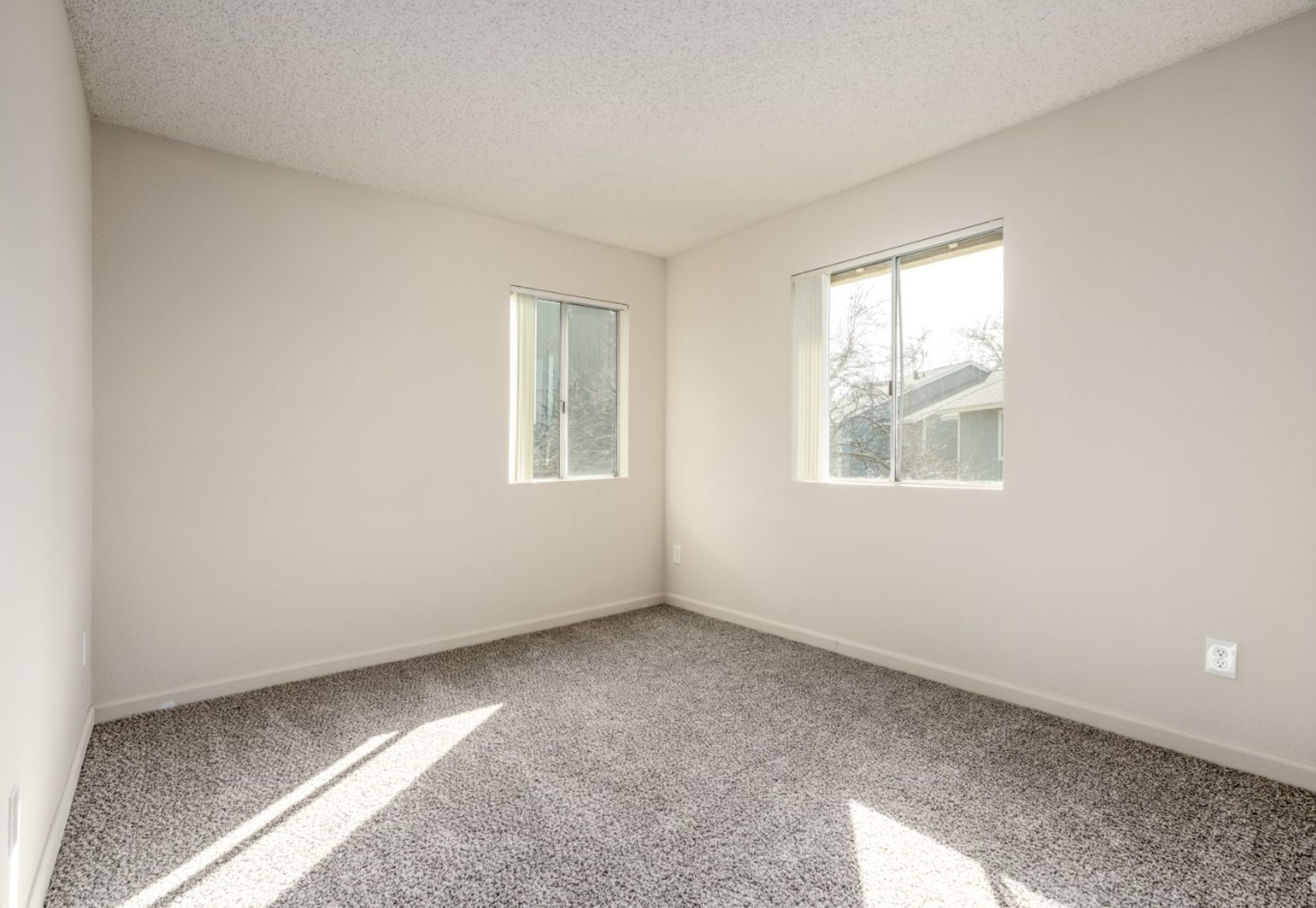 Empty beige bedroom with gray carpet and two windows letting in sunlight