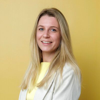 Een close-up portret van een persoon met blond haar, die met een zachte uitdrukking naar de camera kijkt, vlakbij een raam.