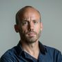 Een persoon met een kaalgeschoren hoofd en lichte stoppels, gekleed in een donkerblauw shirt, kijkt recht vooruit tegen een grijze achtergrond.