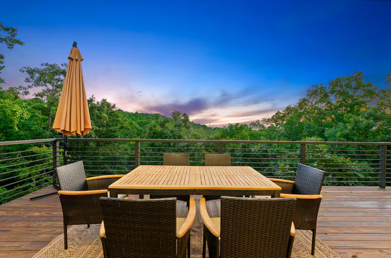 Outdoor dining table and chairs on a wooden deck overlooking lush green trees at sunset.