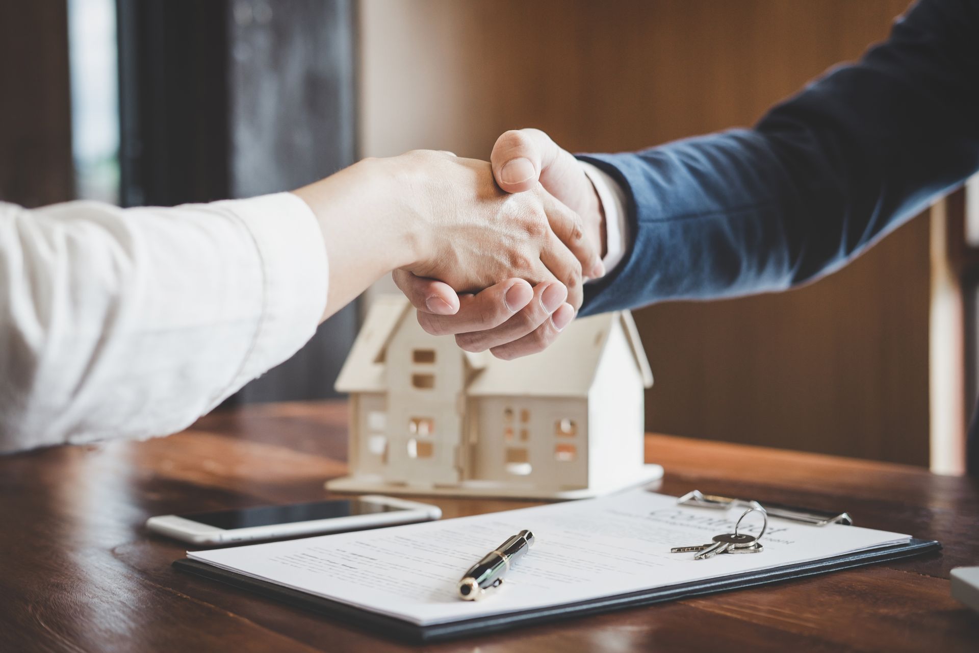 Hands shaking over a real estate contract, small house model, and keys on a table.