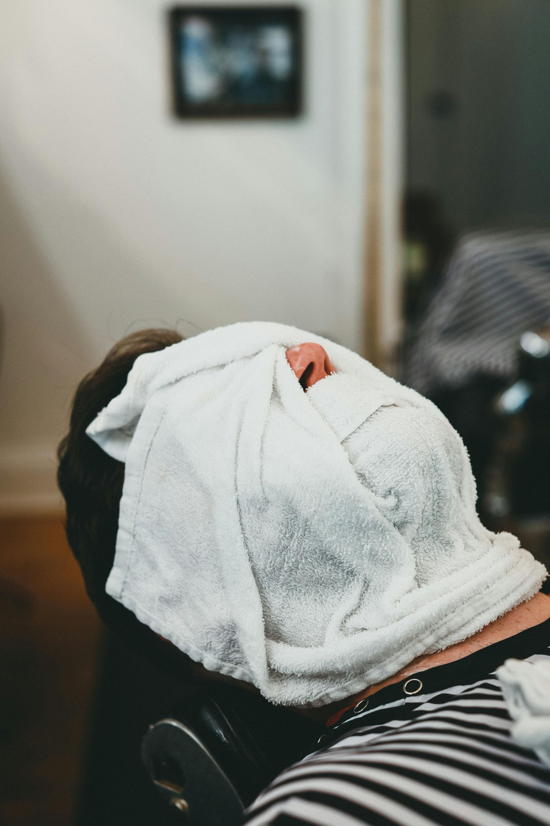 Man with towel over face, getting a shave at a barbershop.