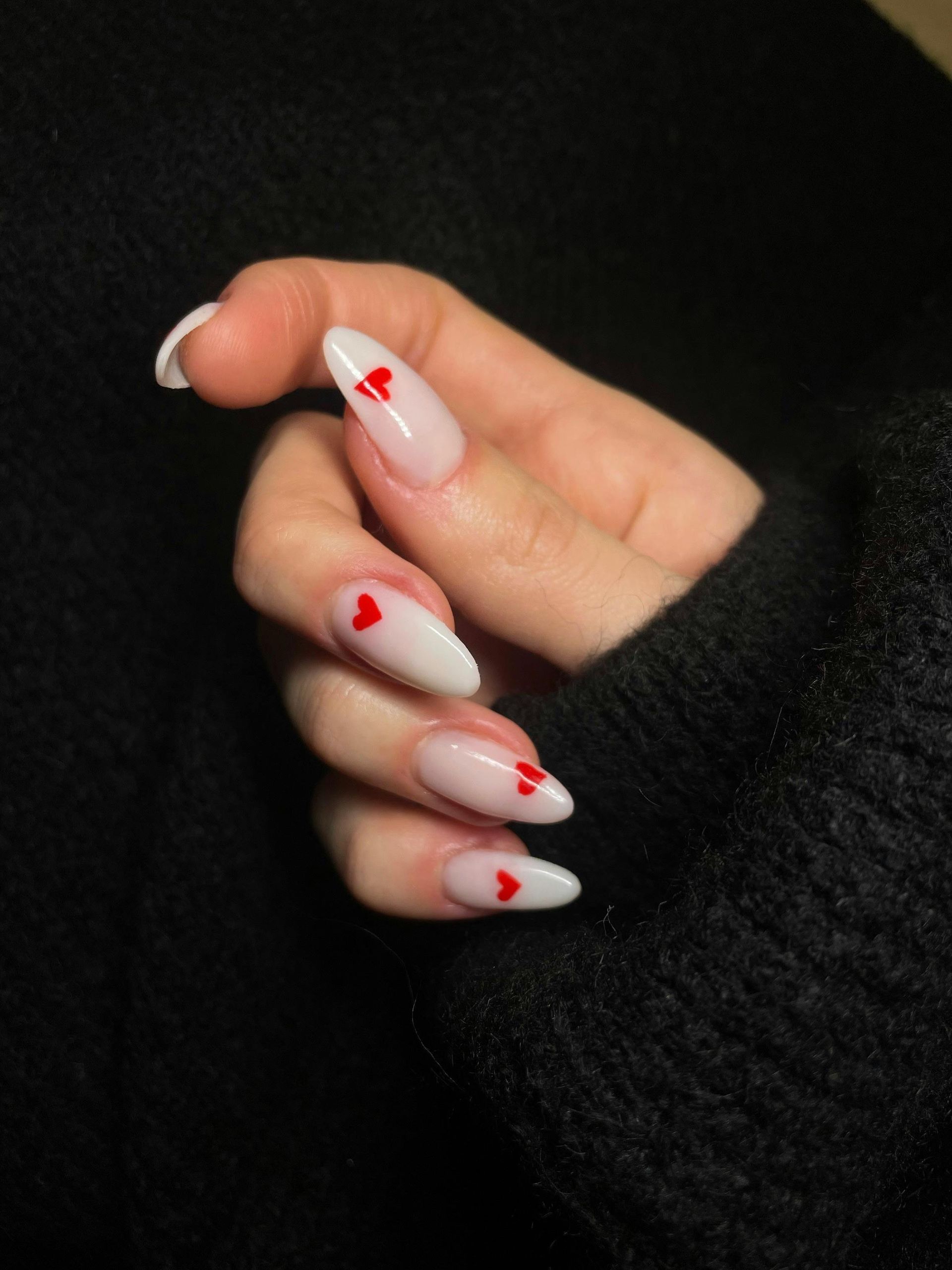 Hand with almond-shaped white nails, each with a small red heart. Dark sweater in background.