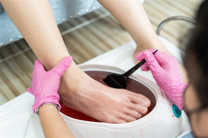 Person getting foot wax treatment at a salon. Two hands in pink gloves, one holding foot, other using spatula.