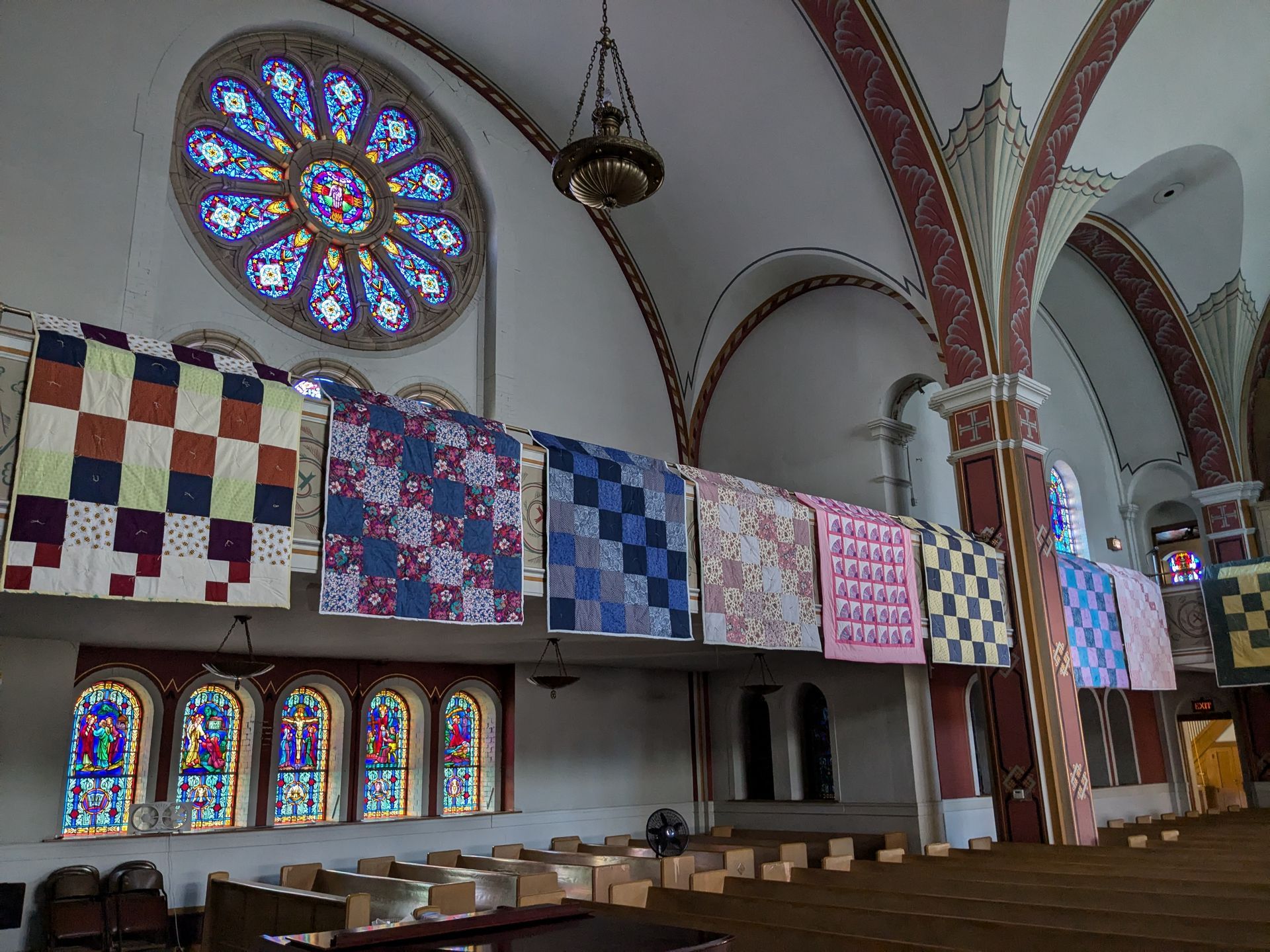 Quilts of various patterns hung inside a church with stained glass windows and arched ceilings.