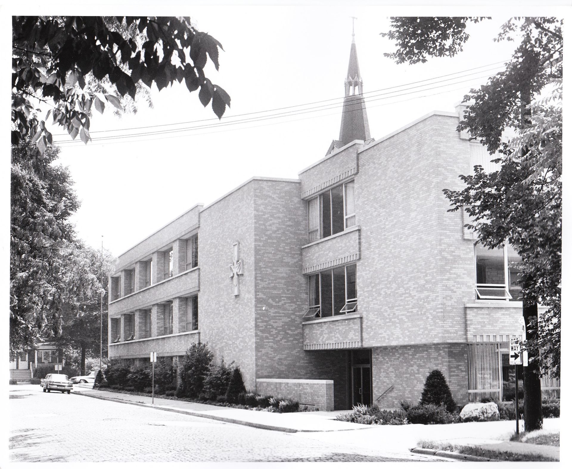 Brick building with a spire, St. John new school building, on a tree-lined street; a car is parked on the road.