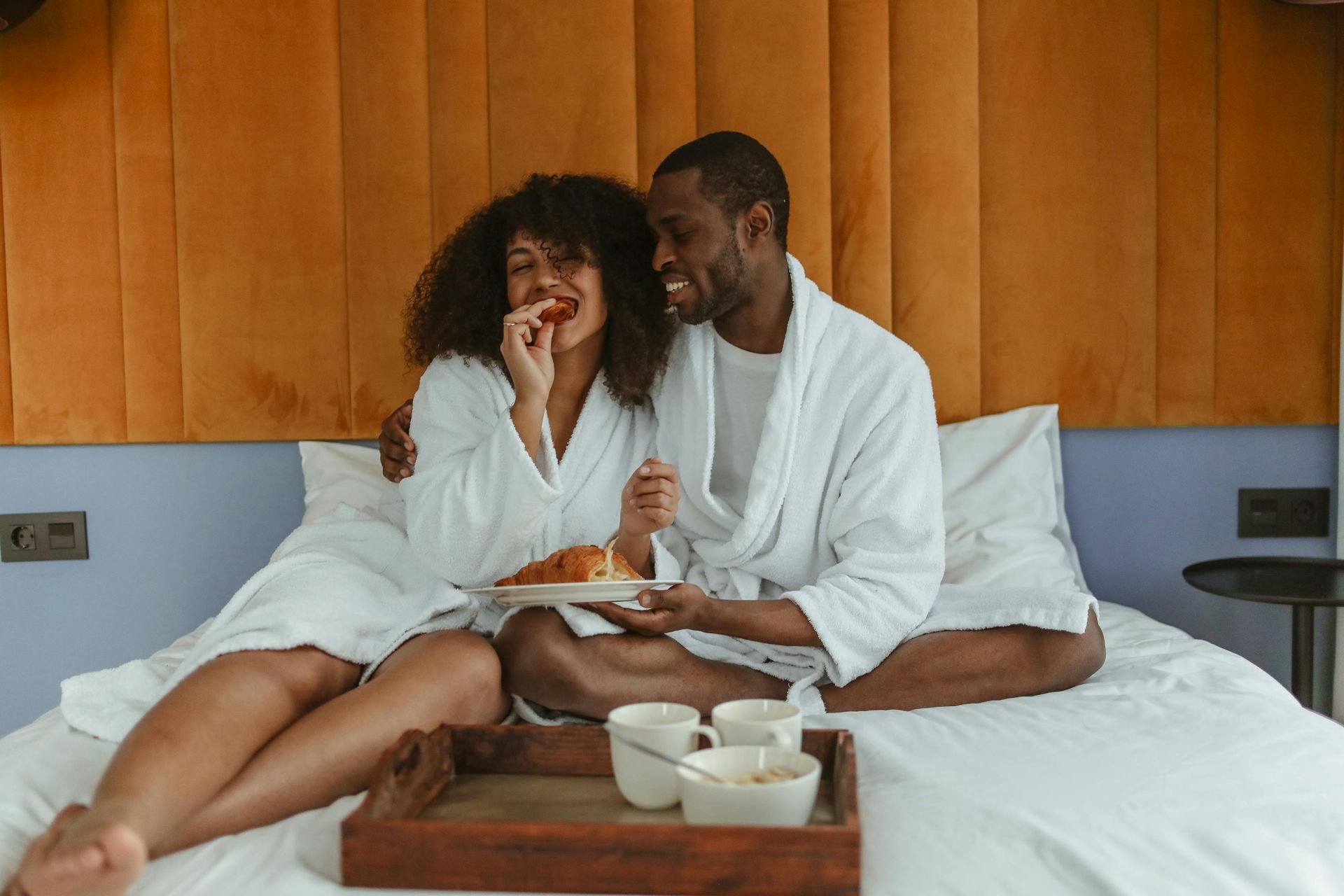 A man and a woman are sitting on a bed with a tray of food.