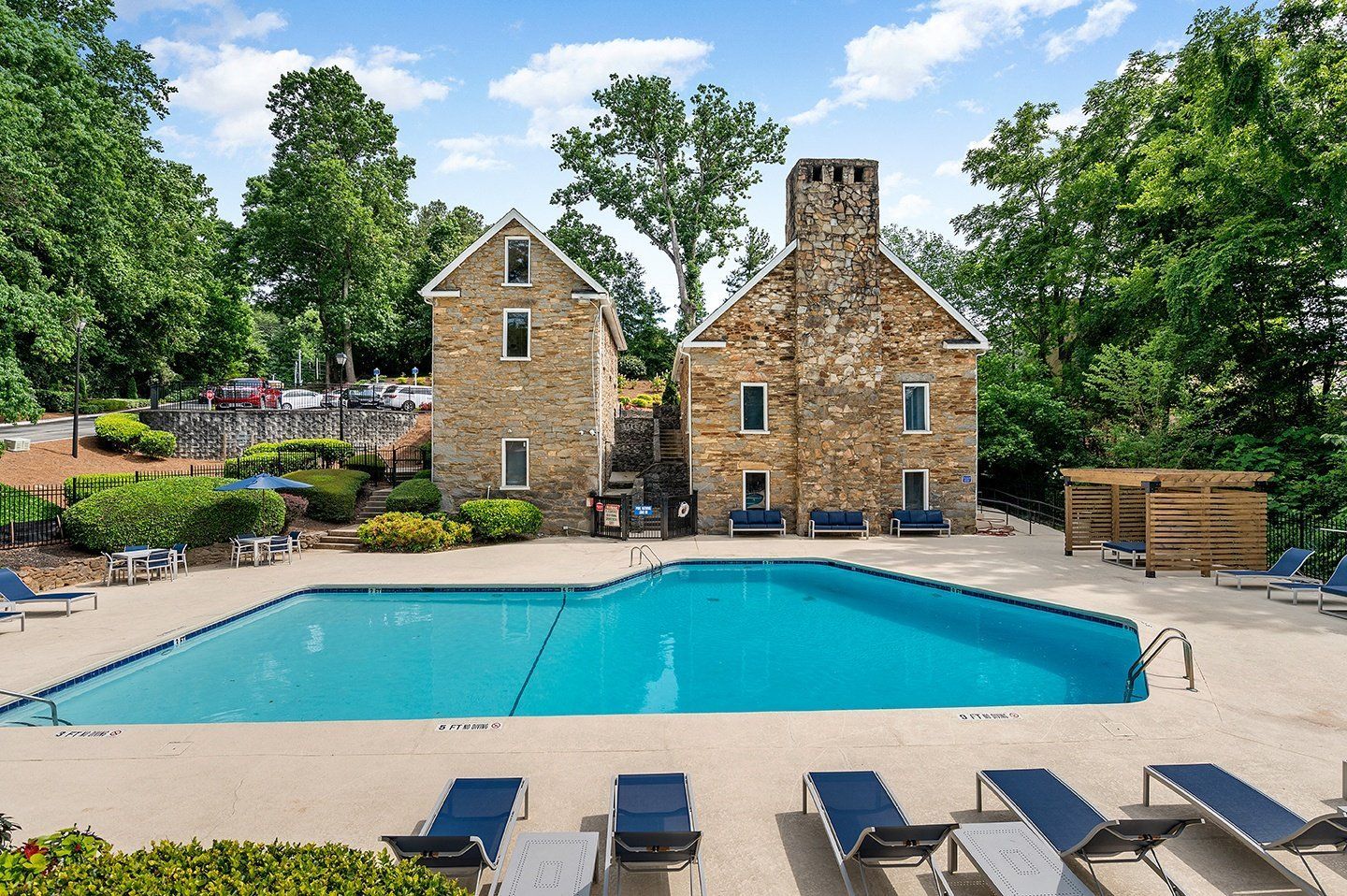 Pool with lounge chairs in front of stone buildings and green trees under a blue sky.