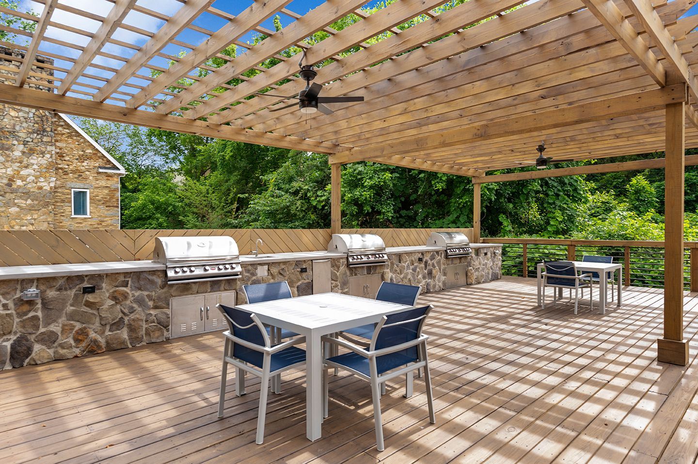 Outdoor grilling area with built-in grills, tables, and a wooden pergola with a fan under a blue sky at Mill House at East Cobb offers apartments for rent in Marietta, GA.