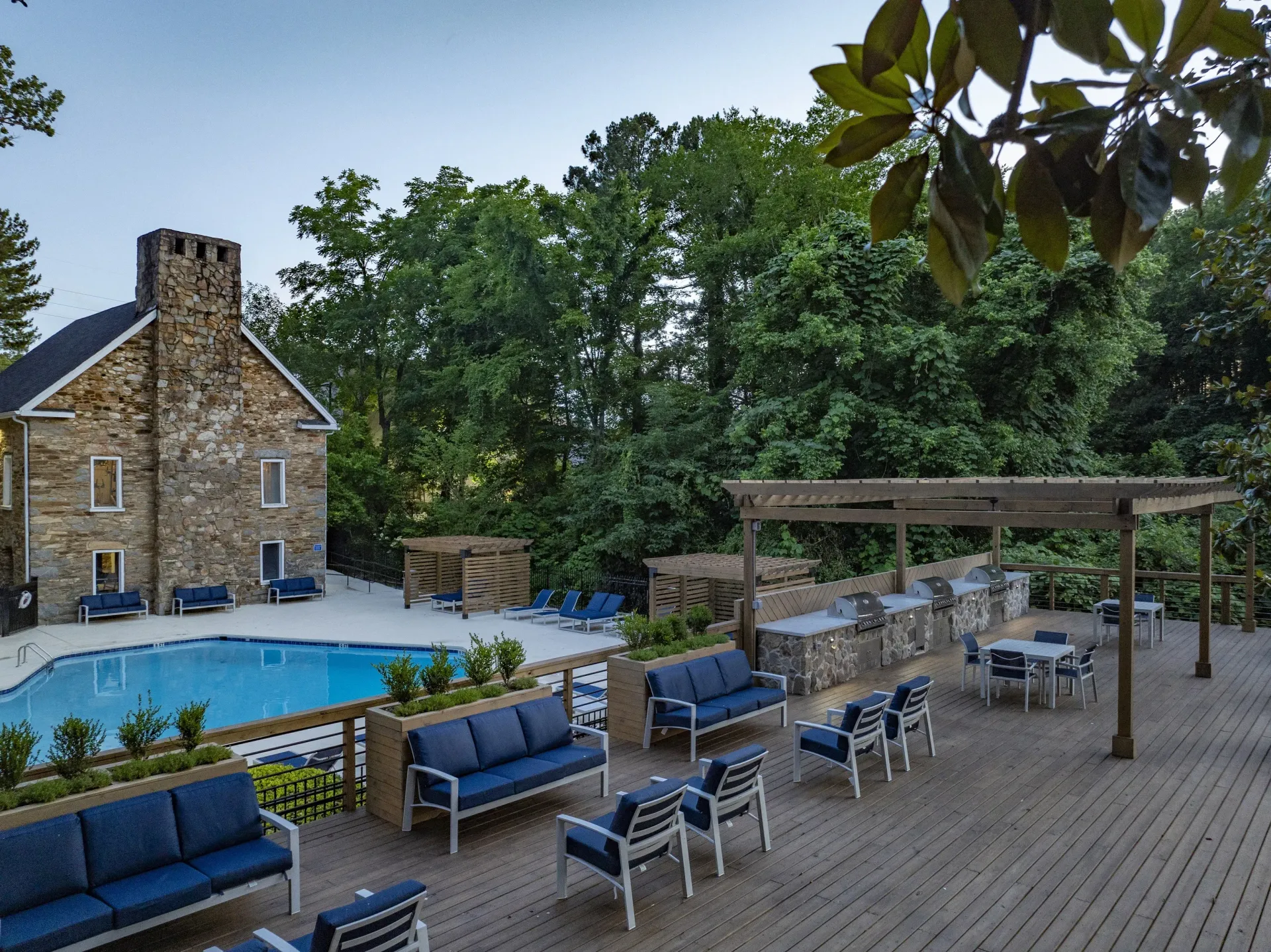 Outdoor pool, lounge area with blue furniture, stone building, surrounded by trees.