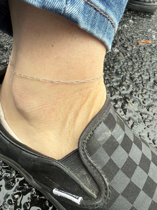 Close-up of an ankle wearing a thin silver anklet and a black shoe with denim jeans above.