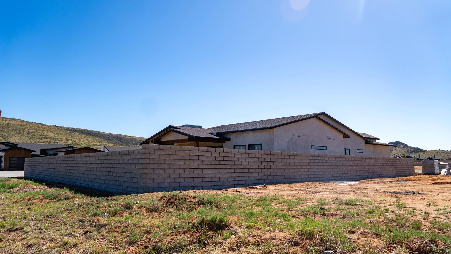 A house with a light-colored facade sits behind a tall, gray brick wall. The sky is blue and the ground is dry with sparse vegetation.