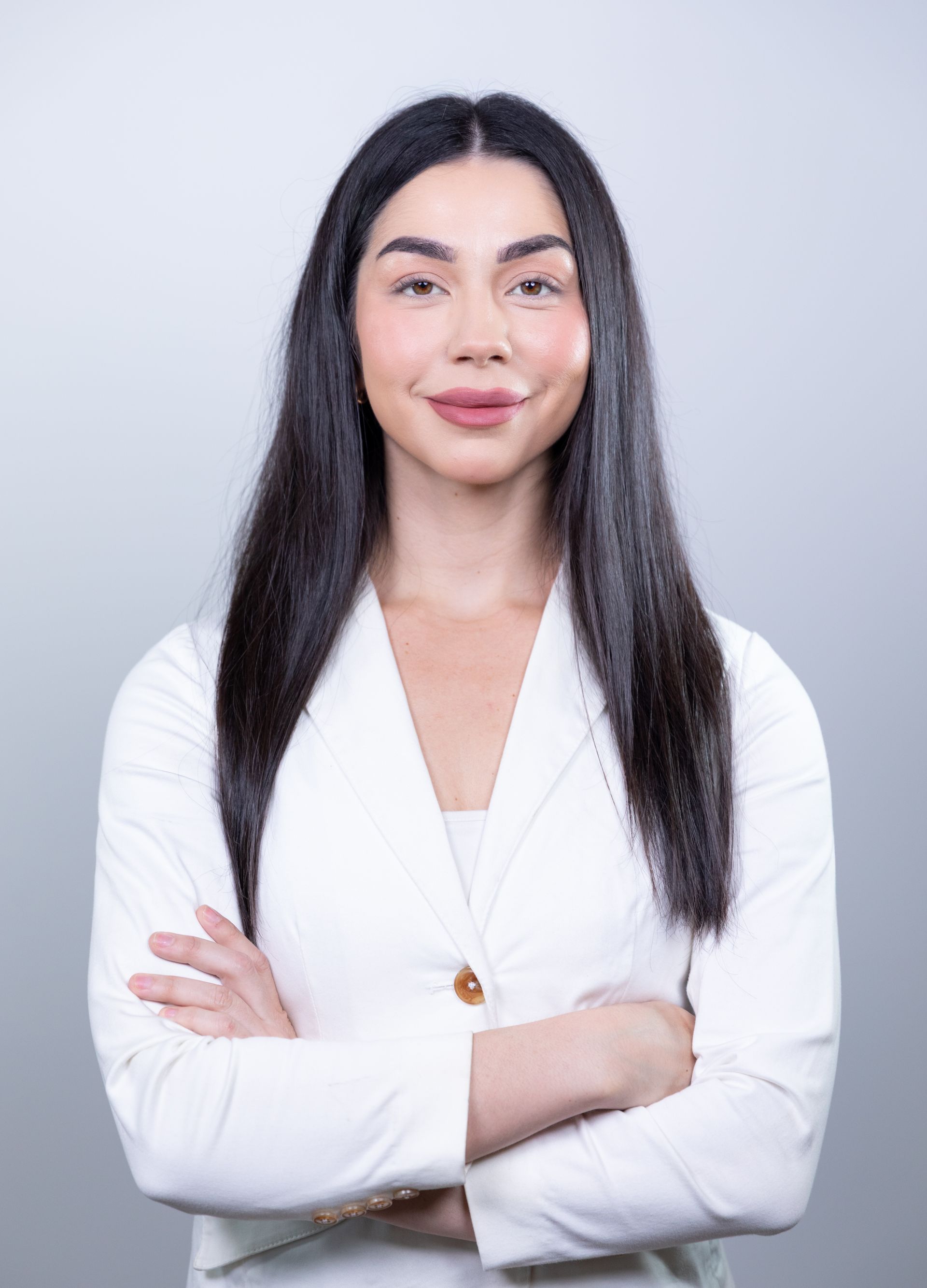 Woman with dark hair in a white blazer, arms crossed, smiling, studio setting.