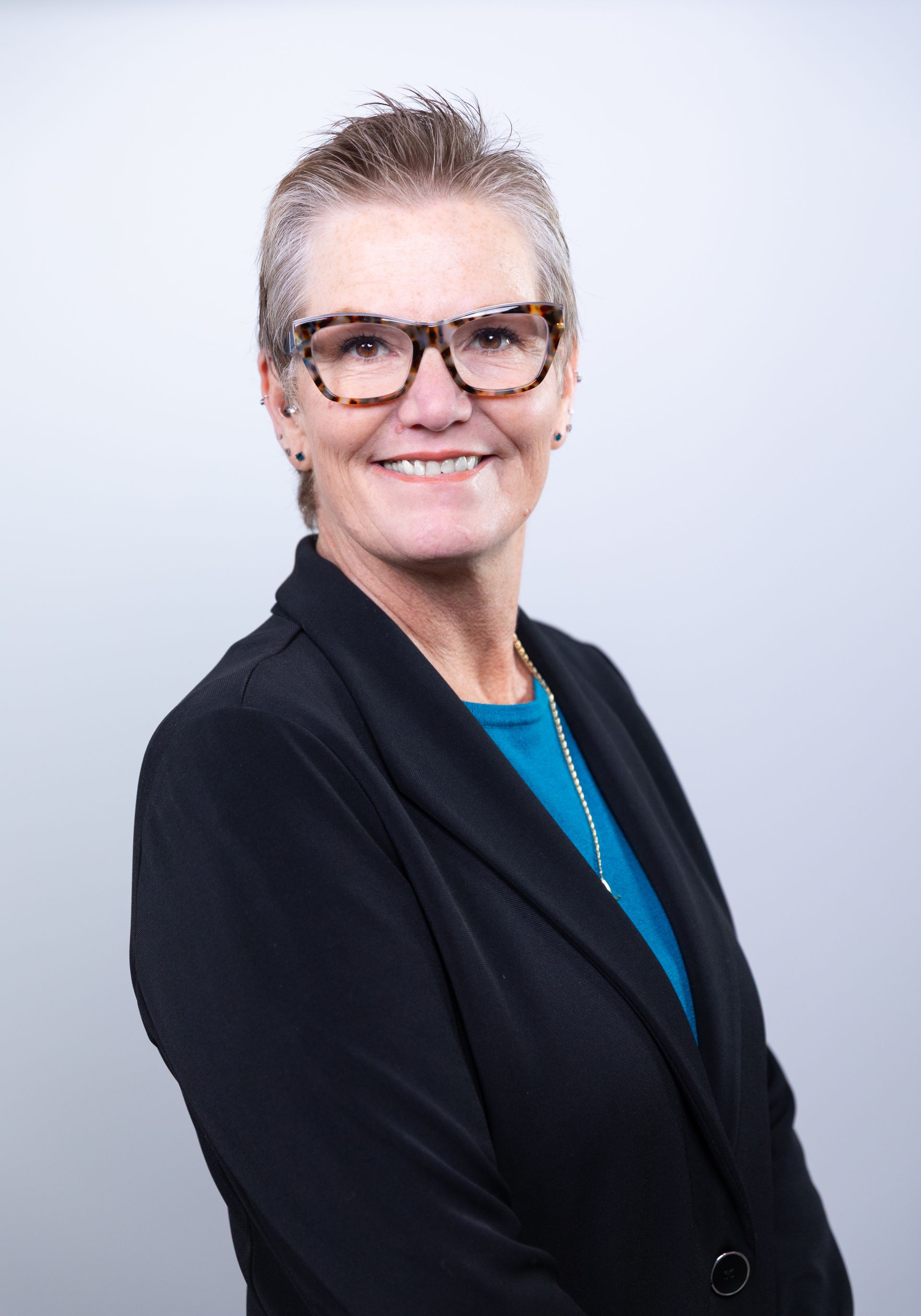 Woman with short hair, wearing glasses and a blazer, smiling, against a light gray background.