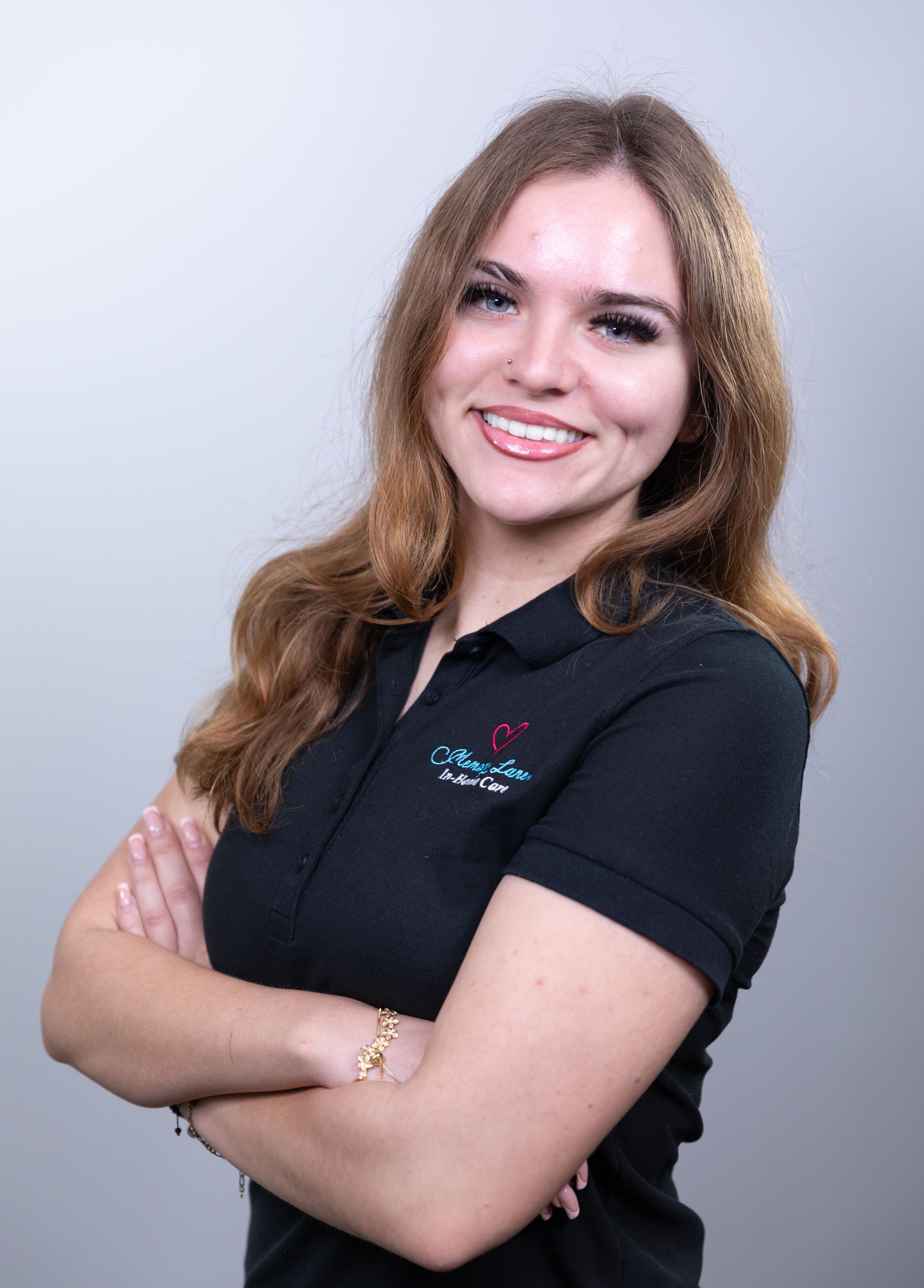 Woman with long brown hair, smiling with arms crossed, wearing a black polo shirt, against a gray background.