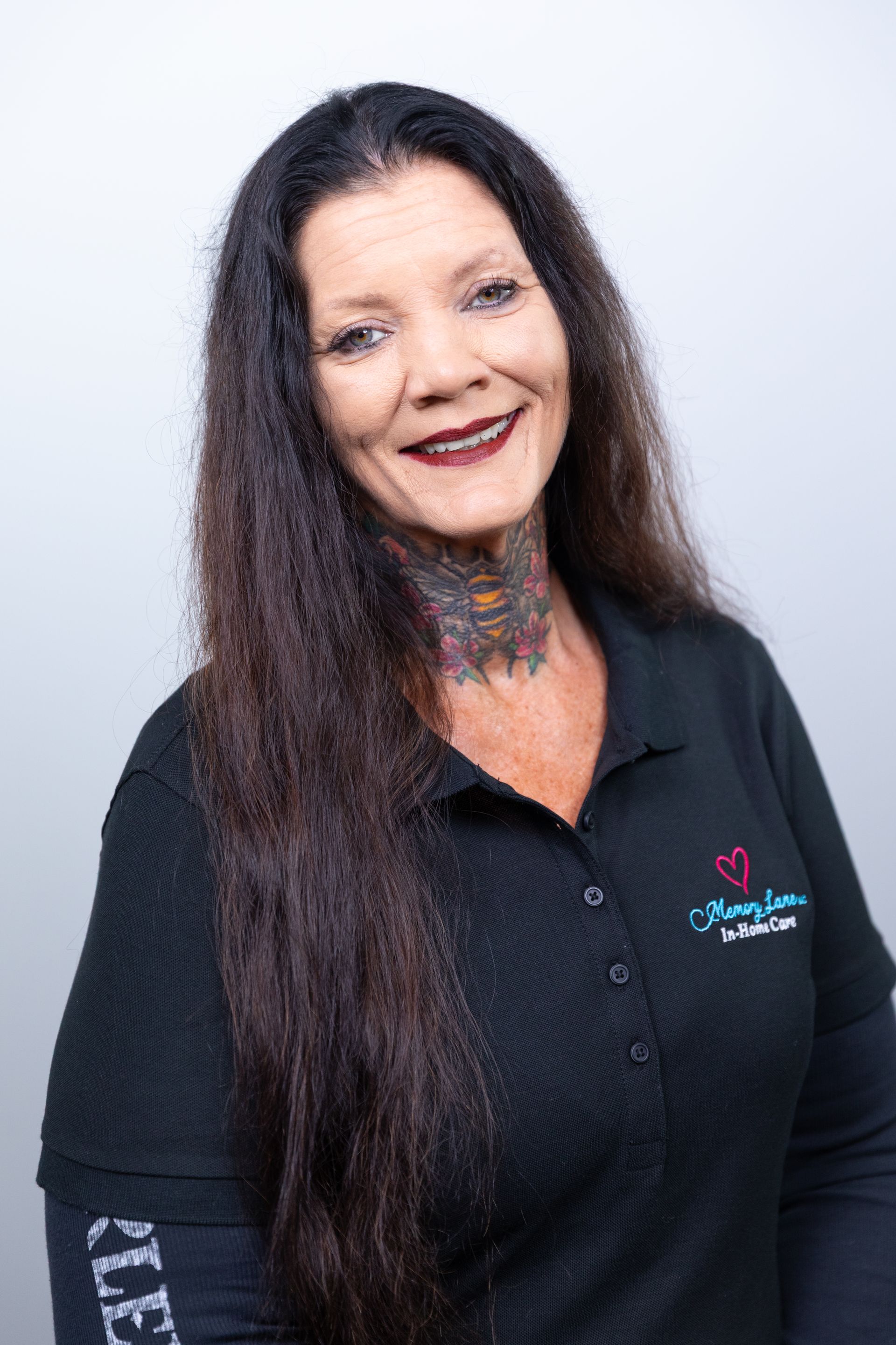 Woman with long dark hair, smiling, wearing a black shirt with a logo and a neck tattoo.