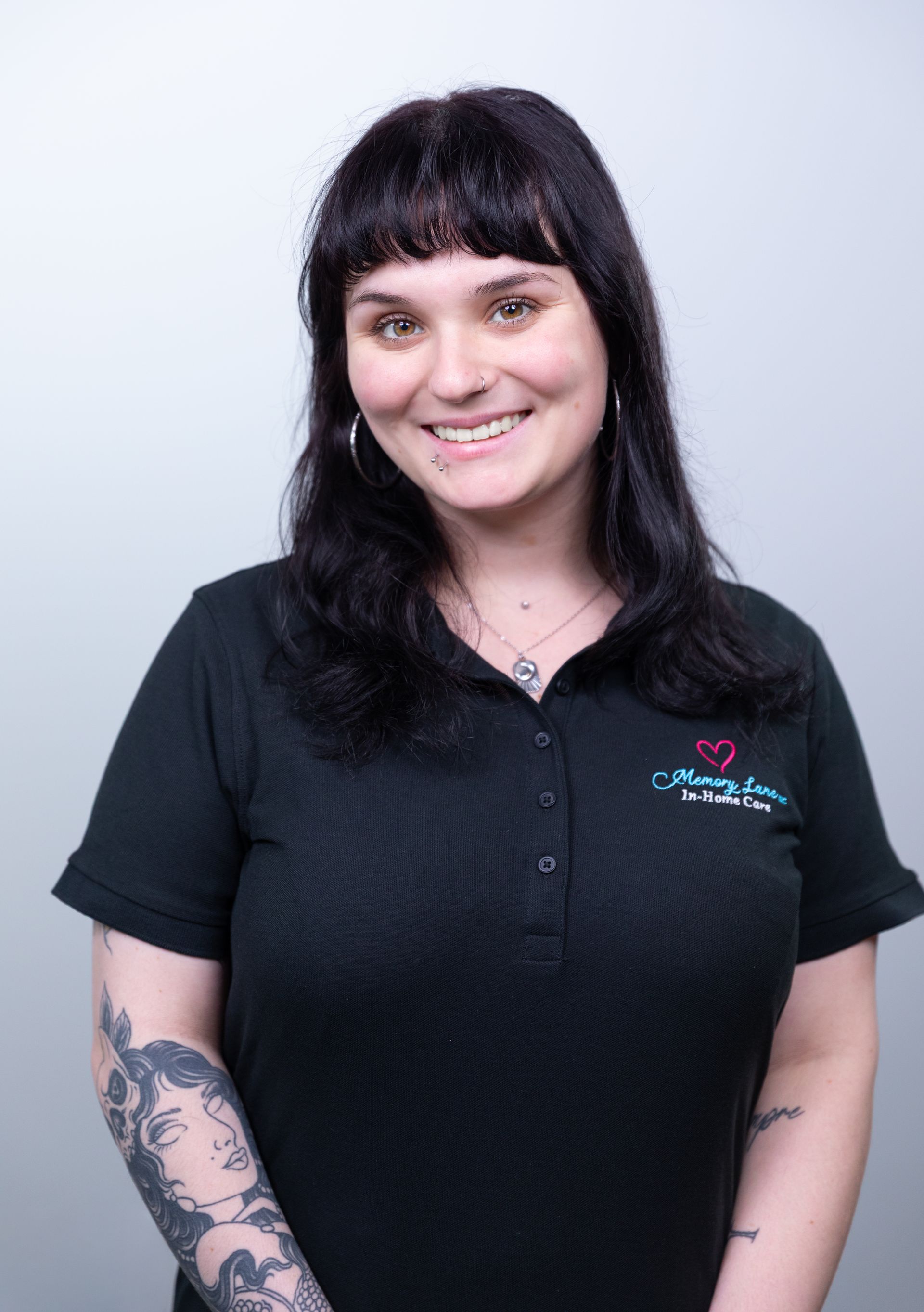 Woman with dark hair smiles, wearing a black shirt with a logo and arm tattoos, stands against a gray backdrop.