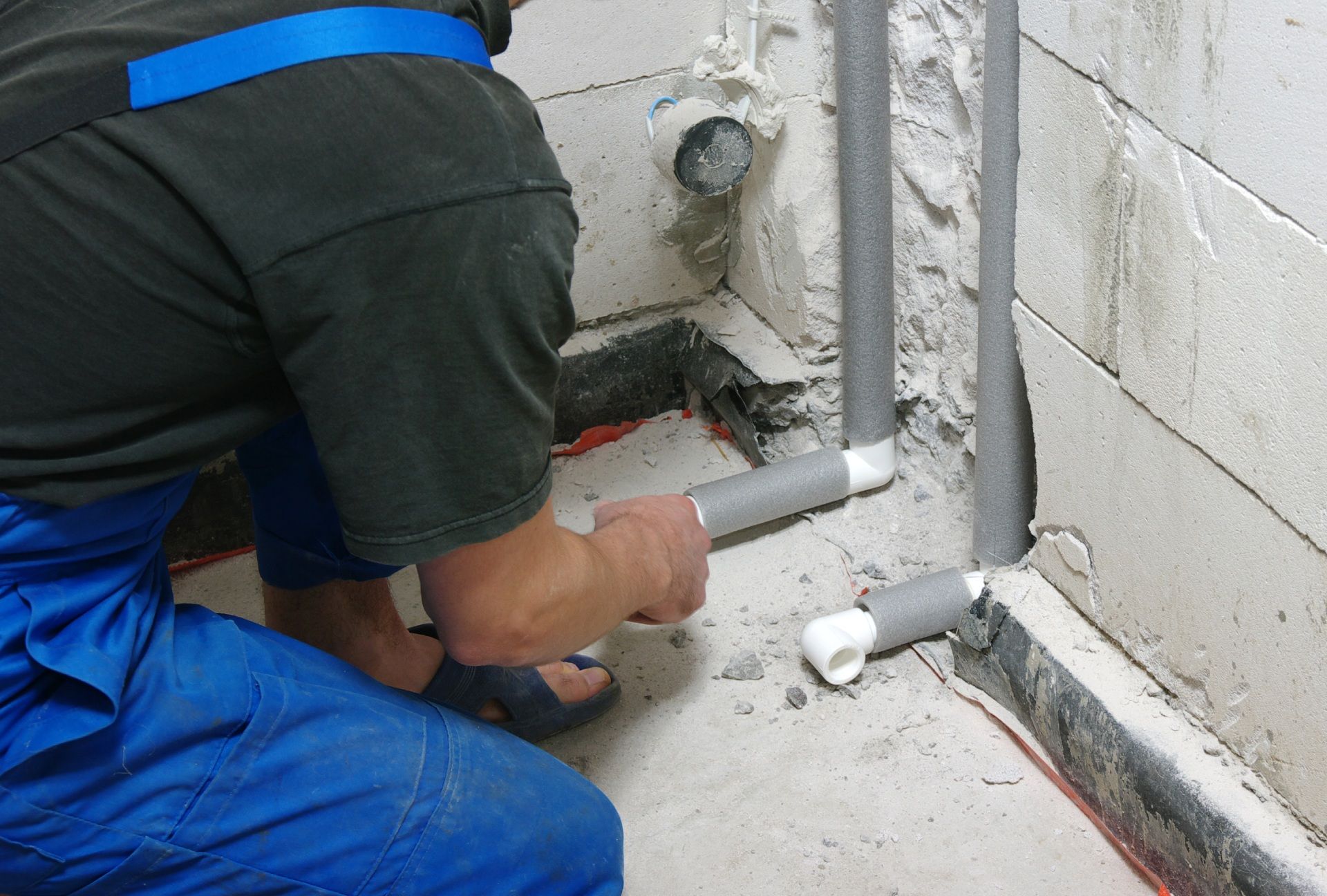 A man in blue overalls is working on pipes in a bathroom.