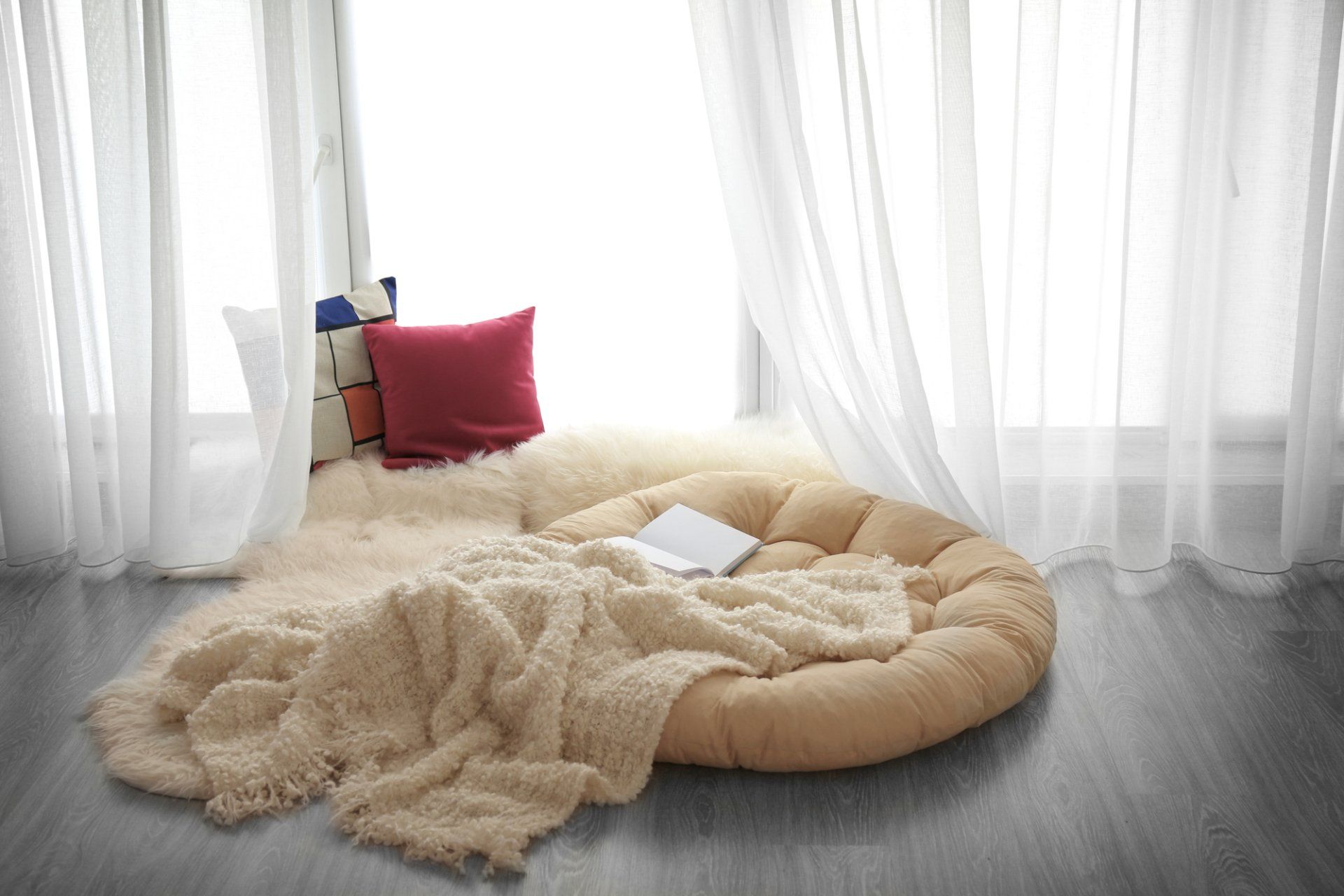 Soft oversize cushion on floor with multiple rugs, backing onto light filled window with opaque sheers.