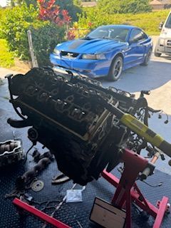 A blue mustang is parked in a driveway next to a engine on a stand.