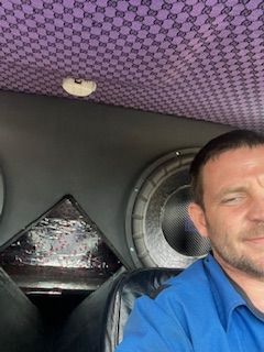 A man in a blue shirt is sitting in a car next to a speaker.