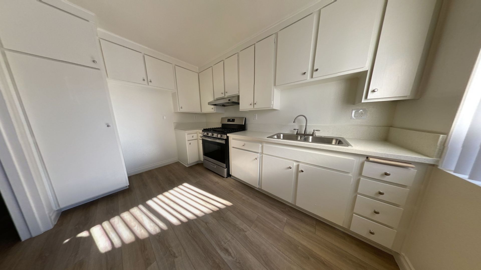 Kitchen with white cabinets, silver appliances, and wood-look flooring. Sunlight streams through a window.