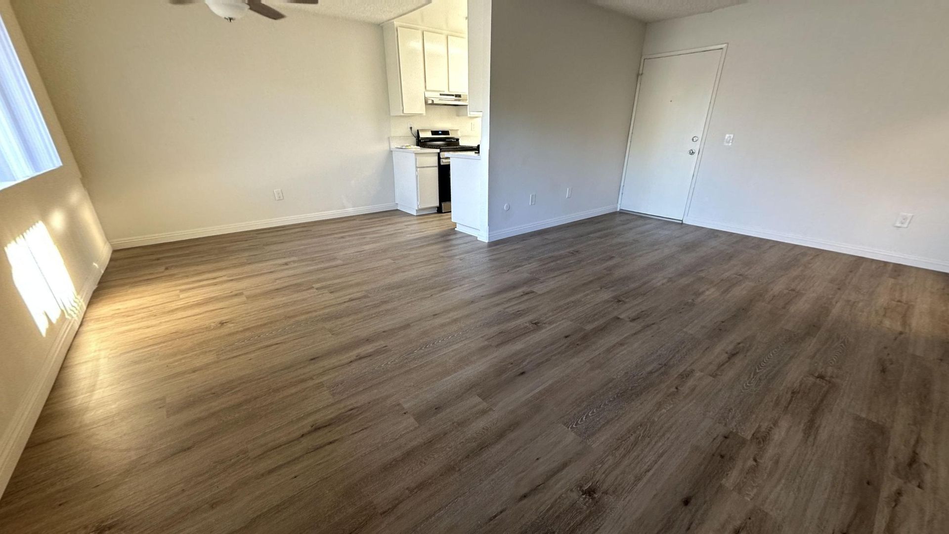 Empty apartment interior with wood-look flooring and white walls, leading to a small kitchen.