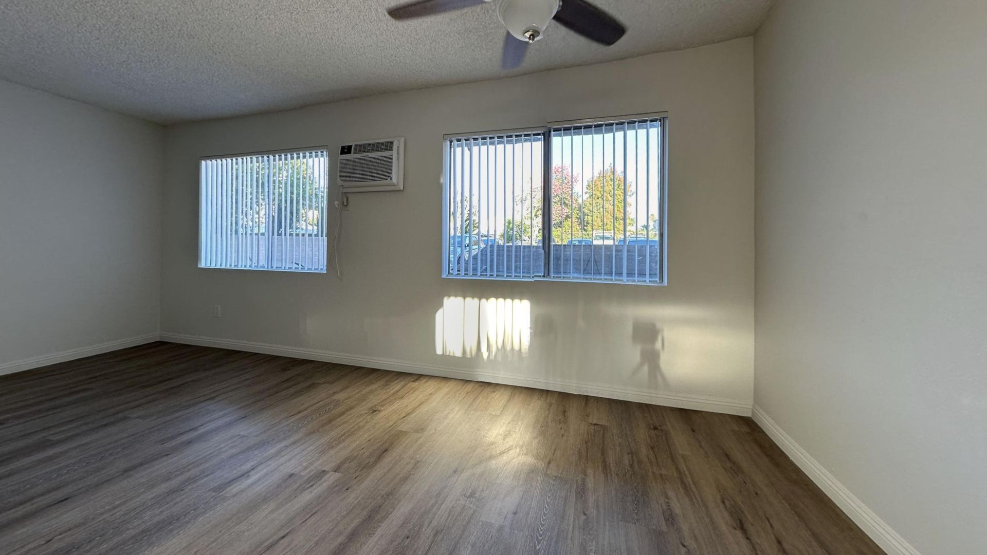 Empty room with wood-look floor, windows with blinds, and ceiling fan. An AC unit is in one window.
