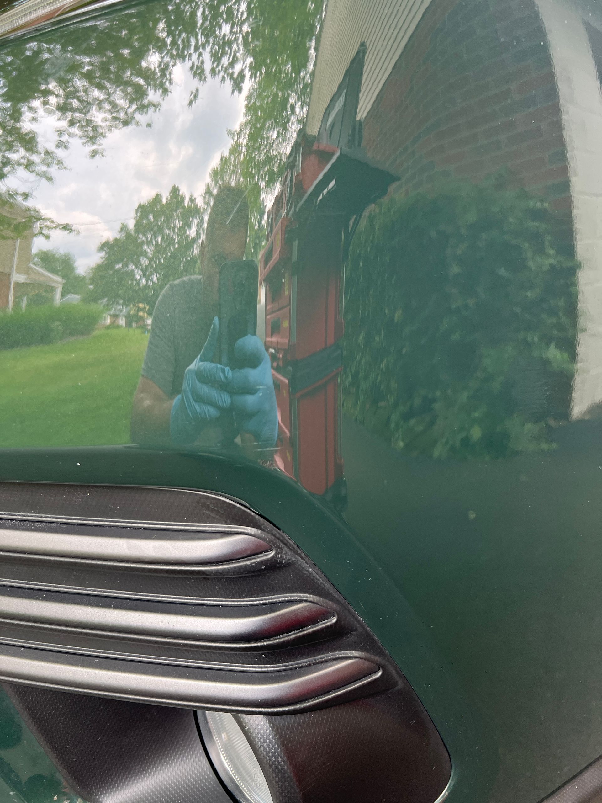 Green car door reflects a person holding an object and a red truck in a residential area.