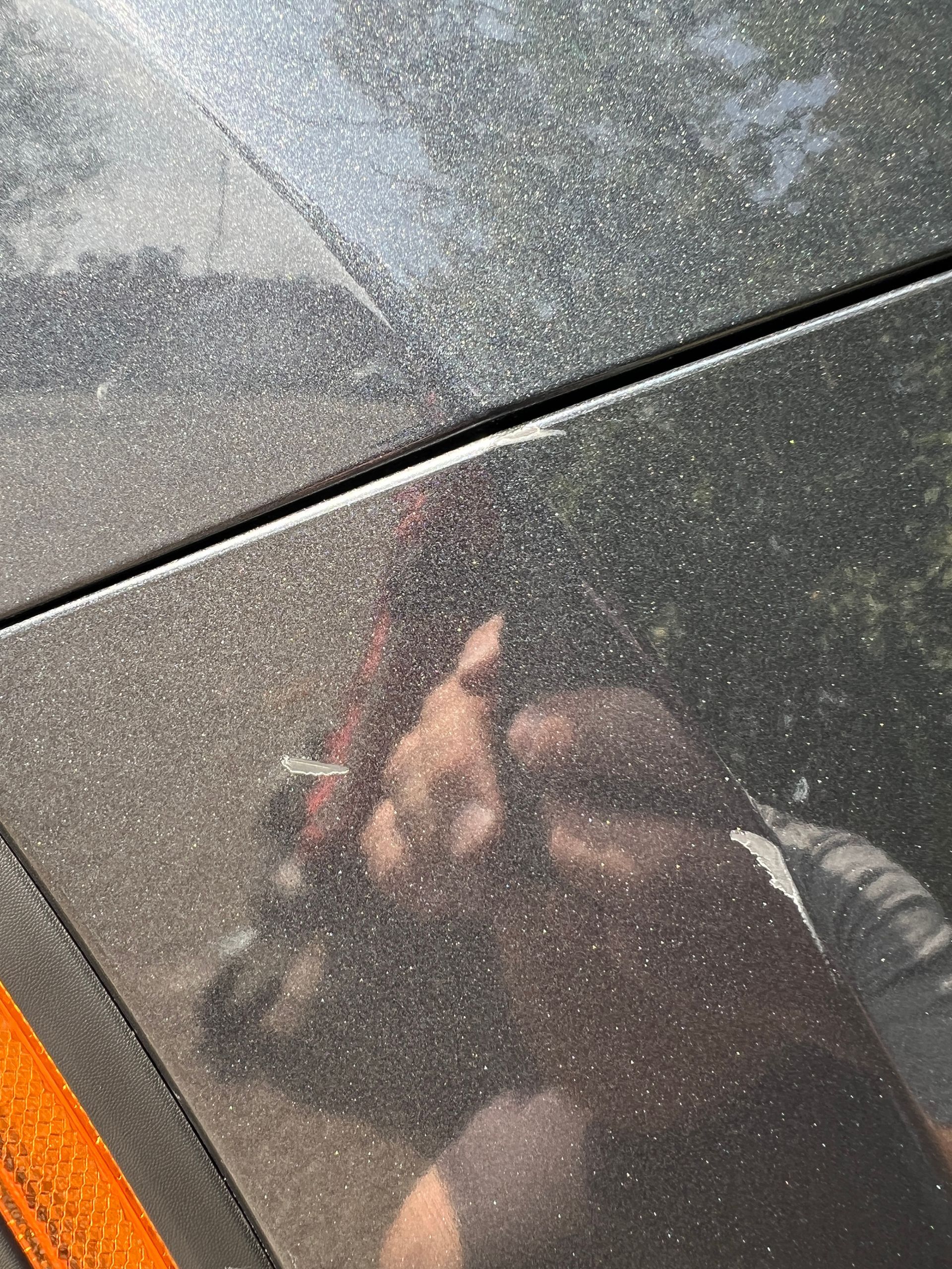 Close-up of a dark, dusty car with a scratch near a panel seam and a person's reflection.