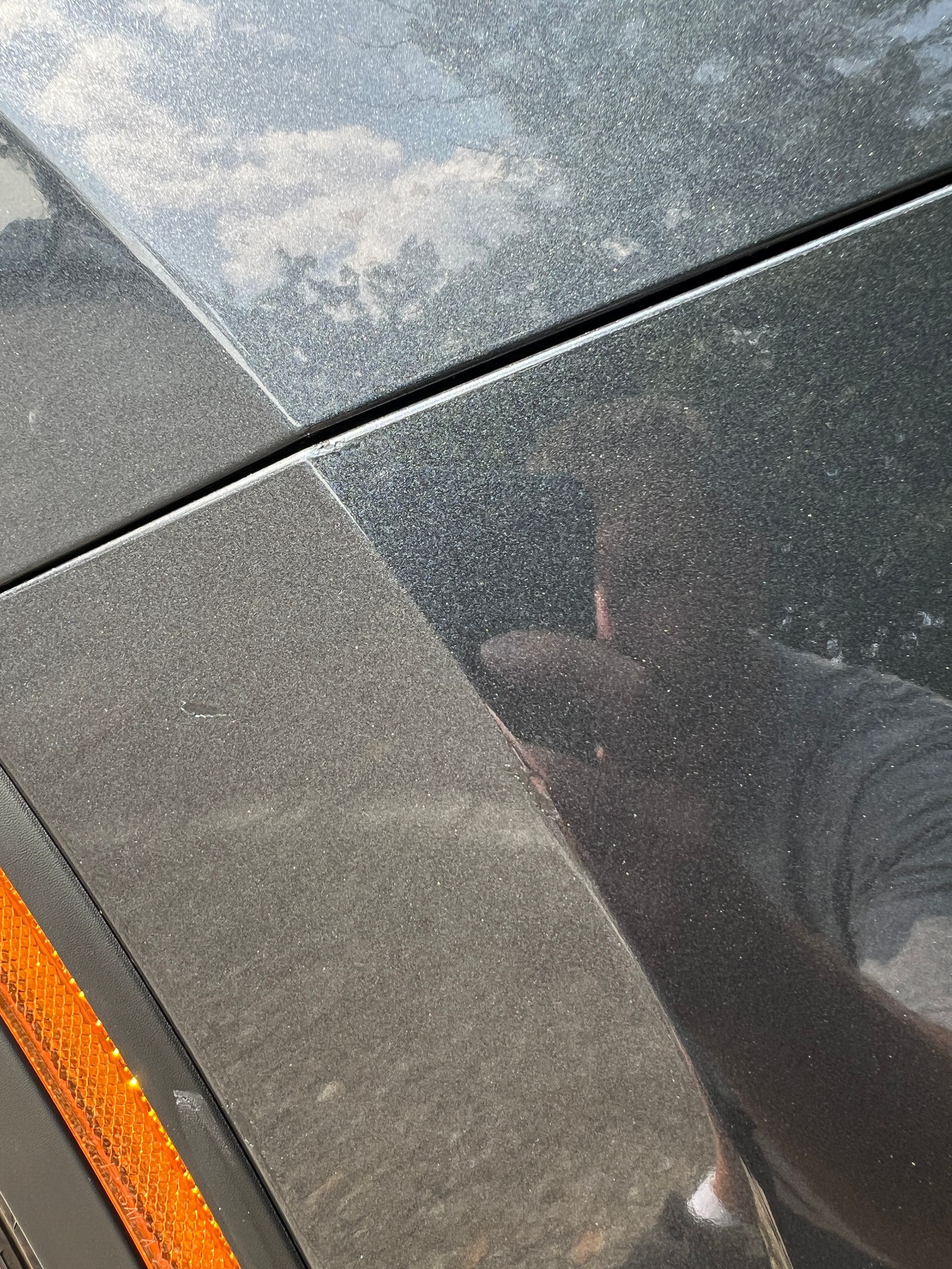 Close-up of a dusty black car with orange side marker light; person reflected on surface.