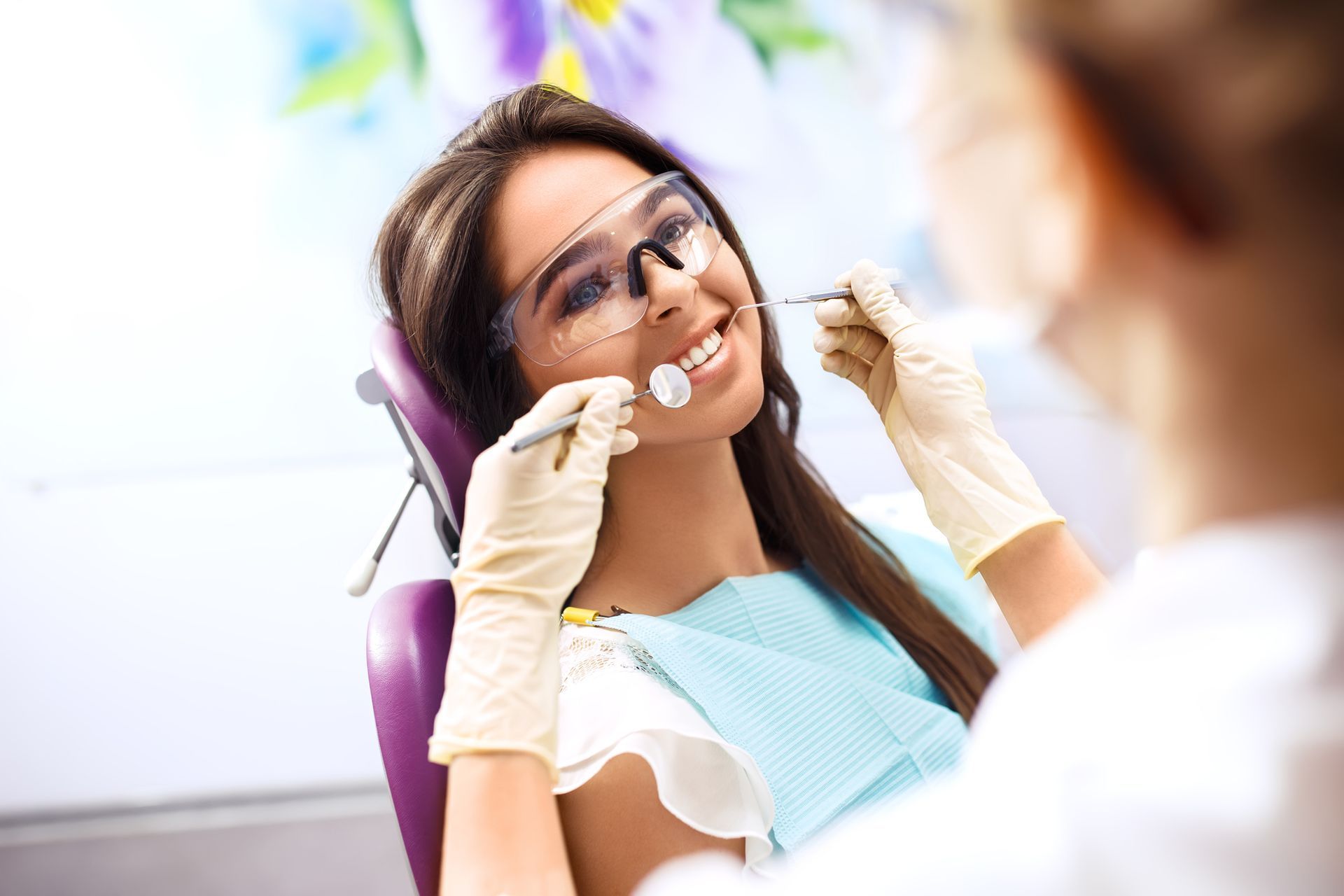 Woman smiling at dentist during a checkup. Wearing protective glasses and bib. Light-colored room.