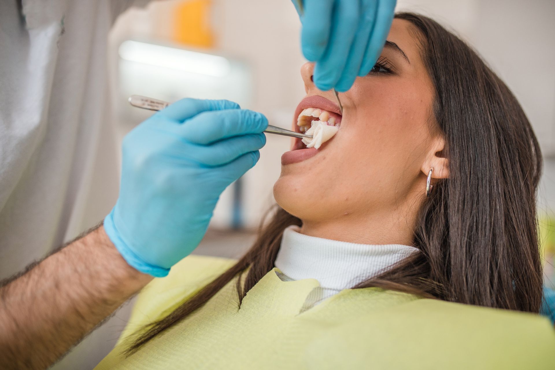 Dentist examining a woman's teeth in a dental chair. The dentist wears blue gloves, and the woman's mouth is open.