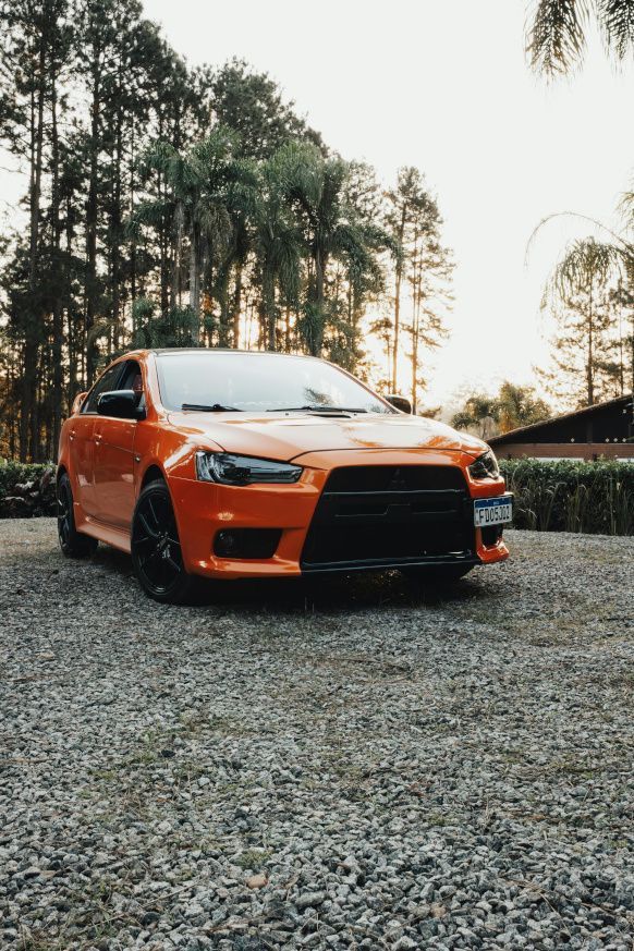 An orange car is parked in a gravel driveway in front of a forest.