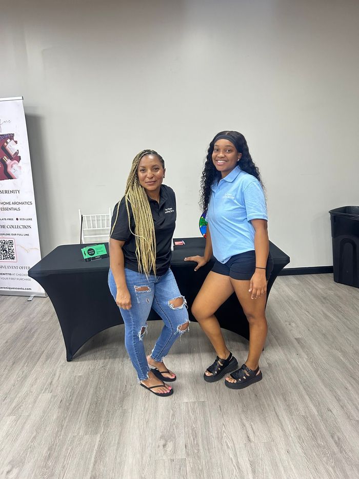 Two women pose by a table. One in jeans and blonde hair, the other in shorts. Inside a room.