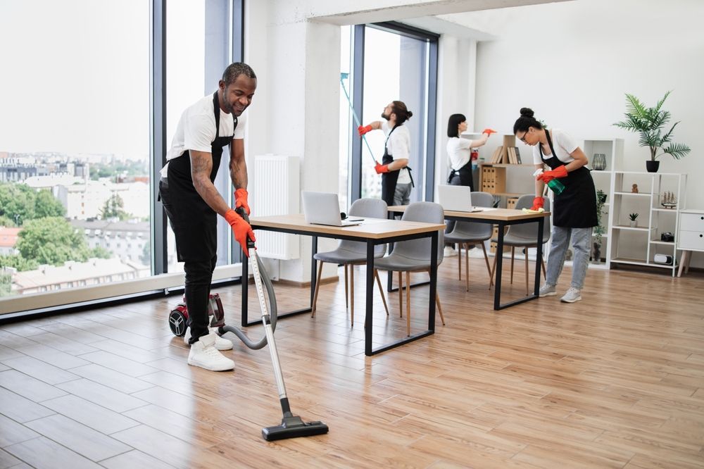 A group of cleaners in an office, vacuuming, wiping windows, and dusting desks.