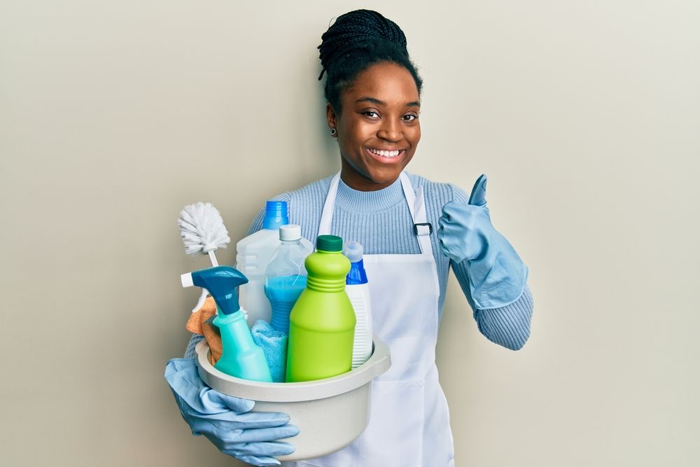 Woman with dark skin in gloves and apron, holding cleaning supplies, giving thumbs up.