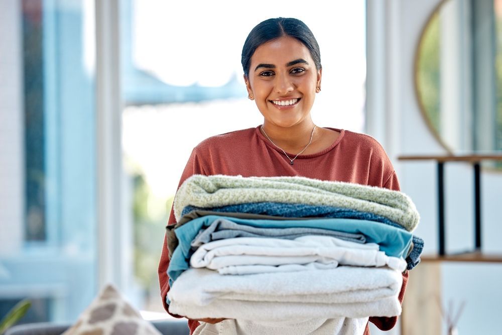 Woman smiling, holding a stack of freshly folded laundry in a bright living room.